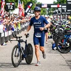 Zawodnik w stroju do triathlonu olimpijskiego biegnie z rowerem obok rzędu rowerów. Tłum kibicuje, trzymając transparent "GO MICHAEL!".