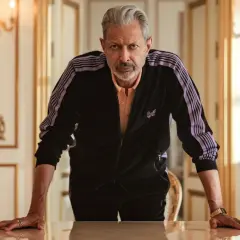 Jeff Goldblum w dresie z fioletowymi pasami, jak z jego filmów, opiera się o stół w eleganckim wnętrzu.