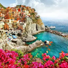 Färgstarka hus klättrar uppför klipporna vid den italienska kusten. Klart turkost vatten och rosa blommor ramar in vyn.