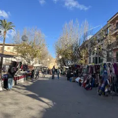 Livlig marknad i Alcudia med stånd som säljer kläder och accessoarer. Palmträd och byggnader kantas av gatan.