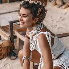 Uśmiechnięta kobieta w stylu boho, ozdobiona muszlami i piórami, siedzi na plaży.
