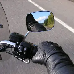 En la carretera, un motorista con guantes de cuero revisa el único espejo de su motocicleta, reflejando el cielo y los árboles.