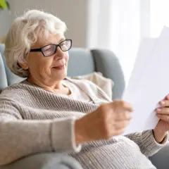 Ältere Frau mit Brille liest Dokumente. Nach einem Herzinfarkt und einer Schwerbehinderung findet sie Trost in der Lektüre.