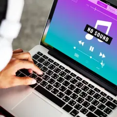 Osoba w słuchawkach próbuje rozwiązać problem **braku dźwięku w laptopie**, na ekranie widnieje komunikat "NO SOUND".