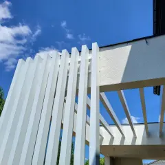 Nowoczesna pergola z białymi, pionowymi lamelami, wykonana z **belki aluminiowe**. Konstrukcja tworzy ciekawy wzór na tle błękitnego nieba z chmurami.