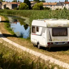 Caravane stationnée au bord d'un canal, dans le Marais Poitevin. Un chemin de terre serpente à travers l'herbe haute.