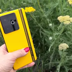 Dłoń trzyma jaskrawożółty telefon z czarnymi paskami, na tle zielonych liści i drobnych żółtych plam kwiatów.