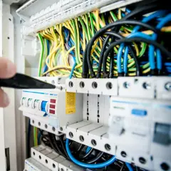 Ręka z narzędziem przyłącze trójfazowe. Widać bezpieczniki i plątaninę kolorowych przewodów w rozdzielnicy elektrycznej.