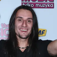 Tomasz Kowalski muzyk z długimi włosami, uśmiechnięty, pokazuje znak pokoju obiema rękami.