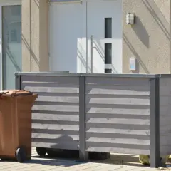 Nowy domek na śmieci to świetny sposób, jak ukryć kosz na śmieci w ogrodzie. Brązowy kosz stoi obok drewnianej obudowy.
