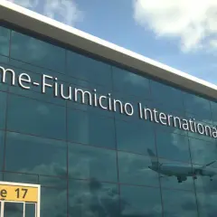 Szklana fasada lotniska w Rzymie, z napisem "Rome-Fiumicino International Airport". W odbiciu widać samolot i chmury.