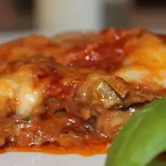 Rebanada de parmigiana di melanzane, con capas de berenjena, salsa de tomate y queso derretido, adornada con una hoja de albahaca fresca.