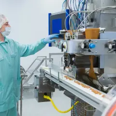 Trabajador en sala blanca, con mascarilla y gorro, supervisa máquina de producción. El asepsia significado se refleja en el control de calidad.