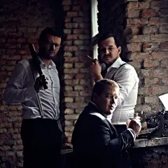 Trzech mężczyzn w białych koszulach i szelkach, jeden z cygarem, obok maszyny do pisania.