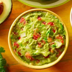 Miseczka guacamole z awokado, pomidorami i cebulą, obok połówka awokado i plasterki jalapeño.
