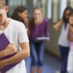Ragazzo solo con quaderno viola, mentre altri bambini ridono e indicano. Sembra avere problemi con maestre elementari.