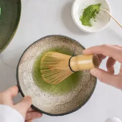 Matcha-Zeit: Hände bereiten grünen Tee mit einem Chasen zu. Daneben ein Stapel grüner Pfannkuchen mit Blaubeeren.