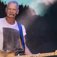 Sting gra na gitarze basowej w białej koszulce na tle sceny.