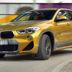 Złoty BMW F39 w ruchu, z widocznym przodem i bokiem, na tle rozmytego miejskiego krajobrazu.