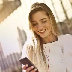 Uśmiechnięta blondynka w białym płaszczu słucha muzyki na telefonie. To inspiracja, jak być kobiecą.