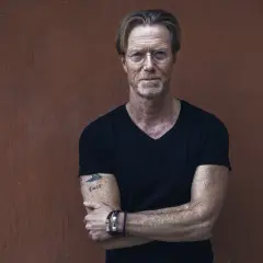 Kommissarie Ewert Grens, en man med intensiv blick och tatuering på armen, står med armarna i kors mot en rödbrun vägg.
