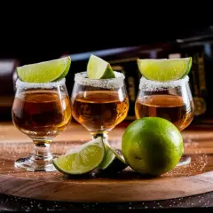 Trzy kieliszki tequili z solą i limonką na drewnianej desce, w tle butelka.