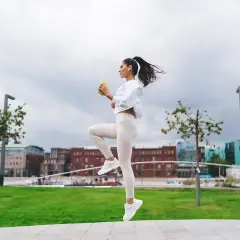 Kobieta w białym stroju sportowym wykonuje dynamiczne ćwiczenie, unosząc kolano. Widać fazy biegu w powietrzu.