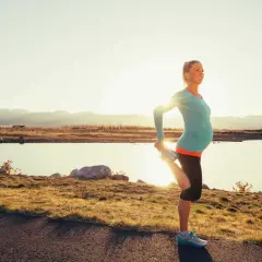 Eine schwangere Frau dehnt sich am Seeufer, genießt das Joggen in der Schwangerschaft bei Sonnenuntergang.
