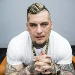 Popek, raper i zawodnik MMA, w białej koszulce i złotym łańcuchu, z tatuażami na rękach i twarzy.