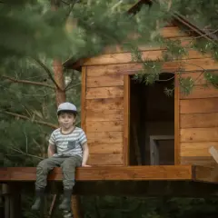 Chłopiec w czapce siedzi na tarasie domku na drzewie. To inspiracja, jak zbudować domek na drzewie dla dzieci.