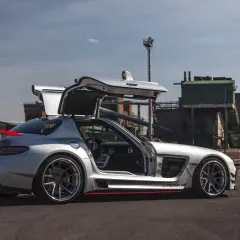 Srebrny SLS z otwartymi drzwiami typu gullwing, z czerwonym akcentem na spojlerze, zaparkowany przed industrialnym budynkiem.