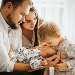 Ciepła sesja noworodkowa z rodzeństwem. Starszy brat delikatnie całuje noworodka, podczas gdy rodzice patrzą z miłością.