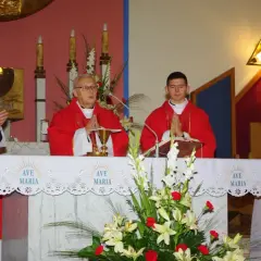 Trzech księży w czerwonych szatach podczas mszy, przed ołtarzem z kwiatami i napisem "Ave Maria".