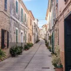 En smal, stenlagd gata i Alcudia old town kantas av gamla hus med gröna fönsterluckor och krukväxter.