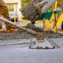 Pracownik wyrównuje świeży beton c12/15 za pomocą łaty. W tle widać koparkę i plac budowy.