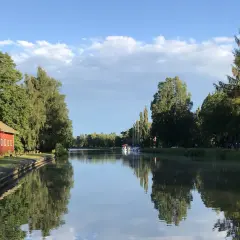 En röd byggnad vid en kanal, troligen Hajstorps slusskafé, med båtar och träd längs vattnet under en blå himmel.