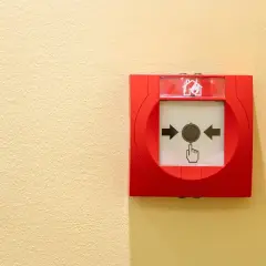 Déclencheur d'alarme incendie rouge sur mur jaune. Une main stylisée pointe le bouton, prêt à signaler une fausse alarme incendie.