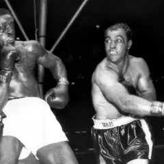 Two boxers, one with white shorts, the other with black, are in the ring. The boxer on the right, with a cut above his eye, is Ezra Charles.