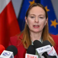 Kobieta w czerwonej marynarce przemawia do mikrofonów. W tle flagi Polski i UE. To po a pis porównanie.