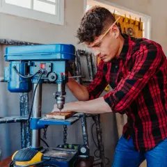 Un homme utilise une perceuse à colonne d'établi pour percer du bois. Il porte une chemise à carreaux rouges et noirs et un crayon derrière l'oreille.
