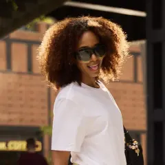 Frau mit lockigem Haar und Sonnenbrille trägt ein weißes T-Shirt.