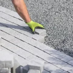 Ręka w zielonej rękawicy układa kostkę brukową na podsypce cementowo piaskowej.