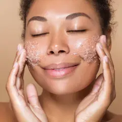 Kobieta z zamkniętymi oczami delikatnie wmasowuje peeling do suchej skóry twarzy. Jej policzki pokryte są drobinkami produktu.