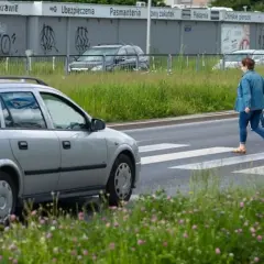 Kobieta w dżinsowej kurtce przechodzi przez przejście dla pieszych. Srebrny samochód czeka, a w tle widać budynki z napisami.
