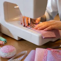 Mains expertes guidant le tissu pour faire des plis précis sur la machine à coudre. Bobines de fil, ciseaux et mètre ruban à proximité.