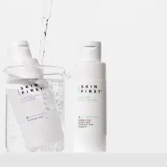Due flaconi di tonico viso Skin First, uno immerso in un becher con acqua e bollicine, l'altro in piedi.