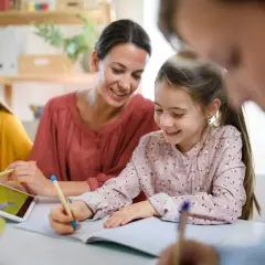 Uśmiechnięta mama pomaga dzieciom w nauce. Jedna córka pisze, druga korzysta z tabletu. To przykład wspólnego uczenia się, gdzie powtarzanie słów, jak w epifora, pomaga zapamiętać.