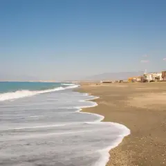 Olas suaves acarician la arena dorada de la playa de Cabo de Gata. Al fondo, un pueblo costero se alza bajo un cielo azul.