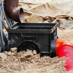 Czarny osadnik rynnowy w kostce, podłączony do czerwonej rury odpływowej, częściowo zasypany ziemią i kamieniami.