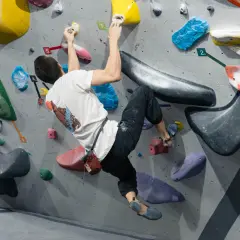Mężczyzna w białej koszulce wykonuje trudny ruch podczas wspinaczki boulderingowej na kolorowej ściance.